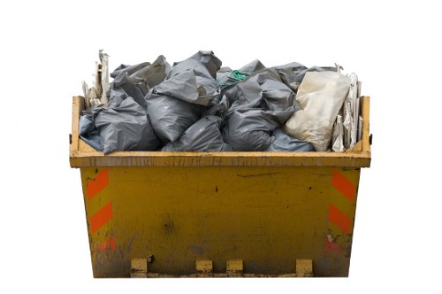 A large yellow metal skip with orange reflective safety markings on its sides, positioned outdoors on a paved surface. The skip is filled to the brim with assorted waste materials, chiefly comprising black plastic rubbish bags and a few lighter-colored items, possibly cardboard or paper. The bags are tightly packed and some are slightly crumpled, with a few extending over the top edge of the skip. The background is plain and white, emphasizing the skip and its contents. This scene is typical of a rubbish removal service in Southwark, where waste management companies like Southwark House Clearance utilize such skips for clearing domestic or commercial waste in the London area. The environment suggests a typical urban setting where builders, homeowners, or tenants might arrange for rubbish clearance. The focus remains on the practical use of the skip for efficient waste disposal, aligned with domestic or construction site rubbish removal in the Southwark district.