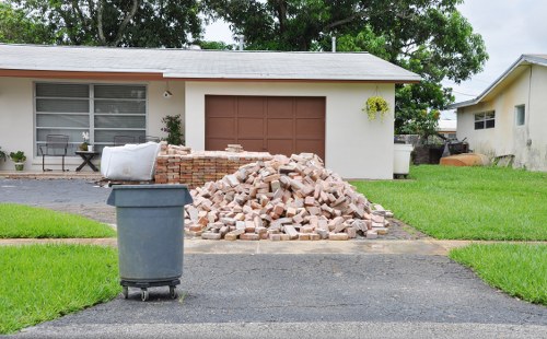 A residential driveway in front of a single-story house with a white exterior and brown garage door, featuring a large pile of beige and light brown bricks loosely stacked on the paved surface. To the left of the brick heap, a dark grey rubbish bin with a closed lid is positioned on the driveway. In the background, the house has a large window with white framing, partially obscured by greenery, and a small shrub growing near the wall. The surrounding lawn is neatly mowed, with vibrant green grass on either side of the driveway, and nearby trees with lush foliage providing a natural backdrop. The scene reflects a typical house clearance process, such as that carried out by Southwark House Clearance, involving the removal of construction debris or renovation waste from a private property in Southwark or nearby areas, with the setting suggesting a quiet suburban neighbourhood.