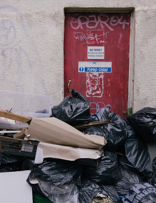 A narrow alleyway between two urban buildings filled with a large pile of mixed waste and discarded materials, including cardboard boxes, plastic bags, and other debris, stacked against a back wall. I