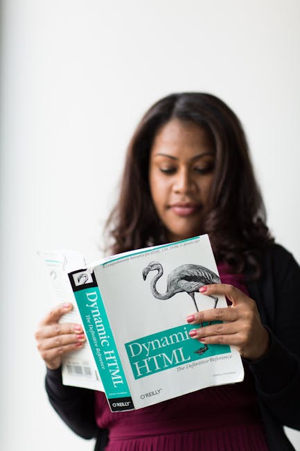A woman with medium-length dark hair is holding and reading a technical book titled 'Dynamic HTML' published by O'Reilly, featuring a black-and-white illustration of a flamingo on the cover, in an ind